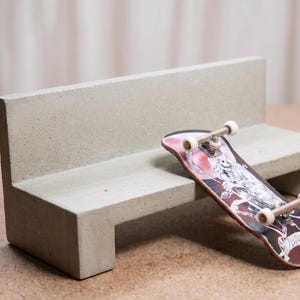 May include: A miniature concrete bench with a smooth, light gray finish and a rectangular backrest. A finger skateboard with a black and red graphic featuring a skeleton illustration and white wheels rests against the front edge of the bench.