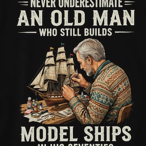 May include: A black graphic t-shirt featuring an illustration of an older person with gray hair wearing a patterned sweater, building a model ship on a wooden desk. The text reads: NEVER UNDERESTIMATE AN OLD MAN WHO STILL BUILDS MODEL SHIPS IN HIS SEVENTIES.