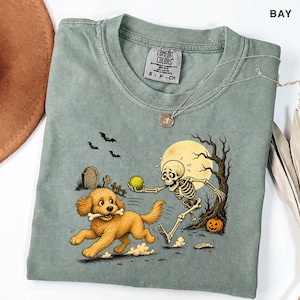 Puede incluir: Una camiseta verde salvia con un gráfico de un esqueleto lanzando una pelota de tenis verde a un perro dorado que sostiene un hueso en la boca. La escena incluye una luna llena, un árbol desnudo, una calabaza de Halloween y murciélagos.