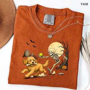 Peut inclure: Un t-shirt orange brûlé avec une illustration représentant un chiot golden retriever tenant un os dans sa gueule, poursuivi par un squelette tenant une balle de tennis verte. Le décor inclut une pleine lune, un arbre dénudé, une citrouille et des tombes.