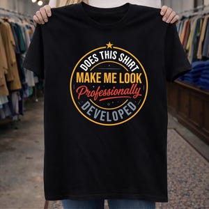 Op de afbeelding: Een zwart t-shirt met korte mouwen en een cirkelvormige afbeelding met een gele ster bovenaan. De tekst "DOES THIS SHIRT MAKE ME LOOK PROFESSIONALLY DEVELOPED" staat in een mix van witte, gele en rode letters met kleine sterretjes.