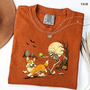 May include: A rust-coloured crew neck t-shirt featuring a graphic of a skeleton playing fetch with a Corgi dog. The scene includes a full moon, a tombstone, a bare tree, and a pumpkin. The shirt has a Comfort Colors brand label at the neck.