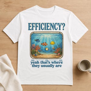 May include: A white short-sleeve t-shirt featuring a graphic of an aquarium with colourful tropical fish, coral, and sand. The text EFFICIENCY? is printed above the aquarium, and the text yeah that's where they usually are is printed below.