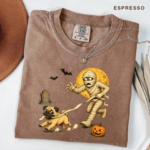 Könnte beinhalten: Ein espressofarbenes T-Shirt mit einer Grafik, die eine Mumie zeigt, die einen Mops an der Leine führt. Das Motiv enthält einen gelben Mond, zwei Fledermäuse, einen Grabstein und einen Kürbis. Das T-Shirt hat einen Rundhalsausschnitt und ein Etikett.