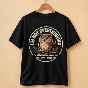 May include: A black short-sleeve t-shirt featuring a circular graphic of a brown owl perched on a branch. The text reads I'M NOT OVERTHINKING and I'M PRE-SOLVING PROBLEMS THAT DON'T EXIST YET. The design includes small mushrooms, stars, and a crescent moon.