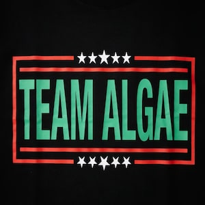 Peut inclure: Un t-shirt noir arborant un motif graphique avec le texte "TEAM ALGAE" en grandes lettres capitales vertes. Le texte est encadré par une bordure rectangulaire rouge, avec cinq étoiles blanches placées au-dessus et au-dessous du bloc central.