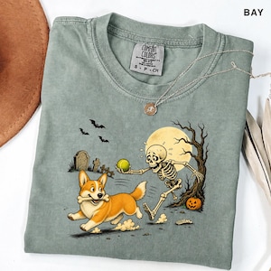 May include: A sage green short-sleeve t-shirt featuring a graphic of a skeleton playing fetch with a Corgi dog. The scene includes a full moon, a bare tree, tombstones, bats, and a jack-o'-lantern. The shirt has a Comfort Colors brand neck label.