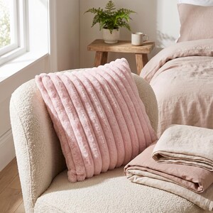 Puede incluir: Un coj&iacute;n cuadrado con una textura de piel sint&eacute;tica de color rosa claro que presenta canales acanalados verticales. El coj&iacute;n est&aacute; colocado sobre un sill&oacute;n texturizado de color crema, con ropa de cama plegada en rosa claro y beige cerca.