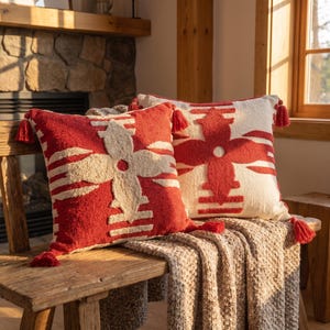 Puede incluir: Dos cojines cuadrados con un patrón floral geométrico con relieve. Uno es principalmente rojo con un diseño en color crema, mientras que el otro es crema con un diseño rojo. Ambos cojines tienen pequeñas borlas rojas en las cuatro esquinas.