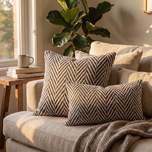 Peut inclure: Deux coussins décoratifs avec un motif à chevrons noir et crème. L'un est carré et l'autre est rectangulaire. Les deux coussins sont disposés sur un canapé beige clair avec un plaid texturé et des coussins unis assortis.