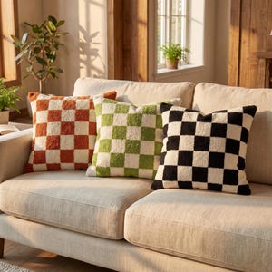 May include: Three square throw pillows with a textured checkerboard pattern. One pillow features orange and cream squares, the second has lime green and cream squares, and the third displays black and cream squares, all arranged on a sofa.
