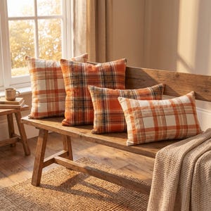Puede incluir: Cuatro cojines rectangulares y cuadrados con un patrón de cuadros en tonos naranja, crema y gris oscuro. Los cojines están dispuestos sobre un banco de madera, mostrando una tela suave y texturizada con un diseño clásico de líneas cruzadas.