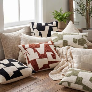 May include: A collection of square and rectangular throw pillows featuring cream-coloured fabric with textured geometric patterns in black, rust orange, and olive green, alongside a solid beige pillow and a cream-coloured chunky knit throw blanket.