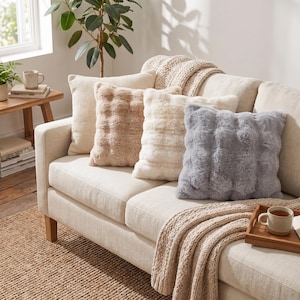 Può includere: Una collezione di quattro cuscini quadrati disposti su un divano di colore chiaro. I cuscini presentano texture varie, tra cui un tessuto color crema a nido d'ape, due cuscini in pelliccia sintetica color beige e bianco sporco, e uno grigio.