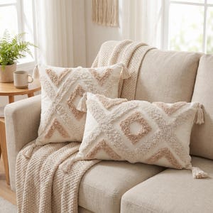 May include: Two off-white throw pillows featuring a textured, tufted geometric diamond pattern in shades of beige and cream. Each pillow is finished with small decorative tassels at the corners, adding a tactile element to the soft fabric design.