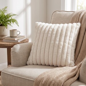 Puede incluir: Un coj&iacute;n cuadrado de color blanco roto con una textura acanalada vertical. El coj&iacute;n tiene un aspecto suave y afelpado similar a la piel sint&eacute;tica y est&aacute; colocado sobre una silla tapizada de color claro con una manta de punto beige texturizada.