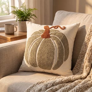May include: A square throw pillow with a cream-colored fabric base featuring a central punch needle design of a pumpkin. The pumpkin is rendered in muted sage green with a textured, raised surface and a prominent rust-orange stem and vine detail.