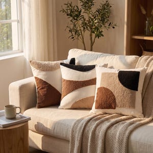 Peut inclure: Trois coussins carrés présentant une base de couleur crème avec des formes géométriques touffetées et texturées dans des tons marron, beige et noir. Les motifs incluent des lignes courbes, des demi-cercles et des blocs abstraits superposés.