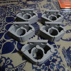 Peut inclure: Cinq supports muraux pour outils en plastique gris clair disposés en groupe. Chaque unité présente un mécanisme de préhension circulaire articulé avec des crêtes internes conçues pour maintenir fermement les manches cylindriques et une patte de fixation plate avec deux trous.
