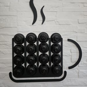 Pode incluir: Um suporte de parede preto para cápsulas de café em forma de uma caneca grande. O suporte apresenta uma estrutura de grade que armazena dezasseis cápsulas de café pretas em quatro filas e quatro colunas, com uma asa curva e volutas de vapor.