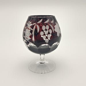 Op de afbeelding: Een dieprood geslepen kristallen cognacglas met gedetailleerde gravures van druiventrossen, bladeren en sterpatronen. Het glas heeft een korte, heldere steel en een ronde voet met een geschulpte rand, wat vakmanschap uitstraalt.