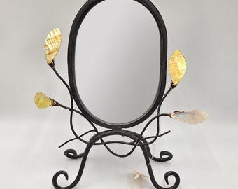 May include: An oval tabletop mirror with a black metal frame and stand. The base features curved legs, and the sides are decorated with thin metal branches adorned with four iridescent, leaf-shaped shell accents in shades of gold and cream.