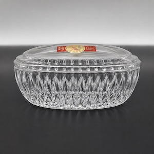 May include: An oval clear glass lidded dish featuring a deep vertical cut pattern along the sides. The lid has a smooth top surface with a small red and gold circular sticker in the center containing the text Cristal d'Arques and 24% PbO.