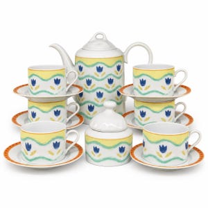Może przedstawiać: Ceramiczny zestaw do herbaty składający się z białego czajnika, cukiernicy z pokrywką oraz sześciu filiżanek z pasującymi spodkami. Każdy element ozdobiony jest powtarzającym się wzorem niebieskich tulipanów, żółtych pasów i falistych zielonych linii.