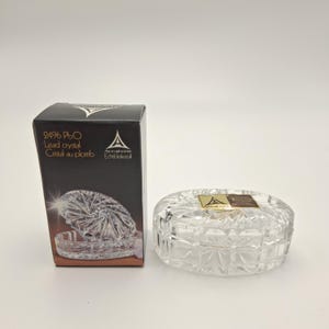 May include: A clear lead crystal trinket box with a faceted geometric pattern and a matching black cardboard box. The packaging features the text 24% PbO, Lead crystal, Cristal au plomb, and the Annahütte logo with the word Echtbleikristall.
