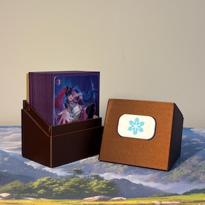 May include: A metallic bronze deck box containing a stack of trading cards with purple sleeves. The front card features an anime-style character with pink hair. The detached lid sits beside the box, displaying a white rectangular emblem with a blue snowflake icon.
