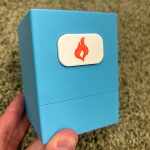 May include: A light blue rectangular deck box with a horizontal white rectangular emblem on the front. The emblem features a centered orange flame icon. The box has a smooth matte finish and a visible horizontal seam separating the lid from the base.