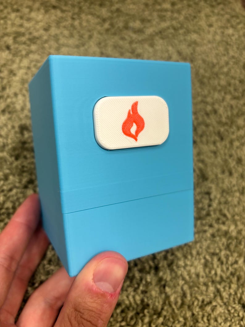 May include: A light blue rectangular deck box with a horizontal white rectangular emblem on the front. The emblem features a centered orange flame icon. The box has a smooth matte finish and a visible horizontal seam separating the lid from the base.