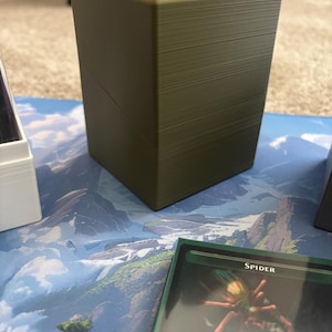 May include: A rectangular olive green plastic deck box with a smooth finish and a diagonal seam across the front. The box features a flat top and vertical sides, designed to hold trading cards, and is positioned near a card labeled Spider.