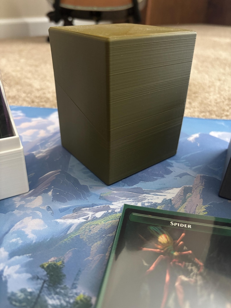 May include: A rectangular olive green plastic deck box with a smooth finish and a diagonal seam across the front. The box features a flat top and vertical sides, designed to hold trading cards, and is positioned near a card labeled Spider.