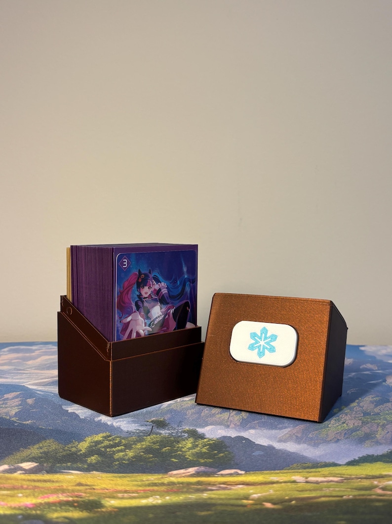 May include: A metallic bronze deck box containing a stack of trading cards with purple sleeves. The front card features an anime-style character with pink hair. The detached lid sits beside the box, displaying a white rectangular emblem with a blue snowflake icon.