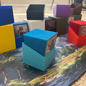 May include: A collection of colorful plastic deck boxes for trading cards, featuring various solid colors including blue, yellow, black, white, purple, brown, red, and light blue. Each box has an angled open top to display the card inside.