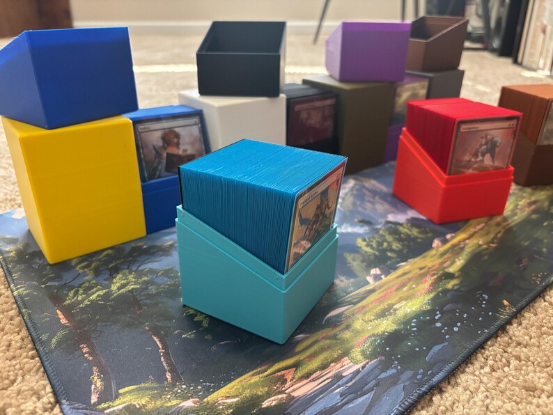 May include: A collection of colorful plastic deck boxes for trading cards, featuring various solid colors including blue, yellow, black, white, purple, brown, red, and light blue. Each box has an angled open top to display the card inside.