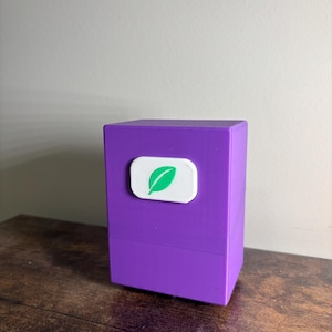 May include: A rectangular purple storage box with a smooth matte finish. Centered on the front face is a white, rounded rectangular emblem featuring a green leaf icon. The box has a horizontal seam line separating the lid from the base section.