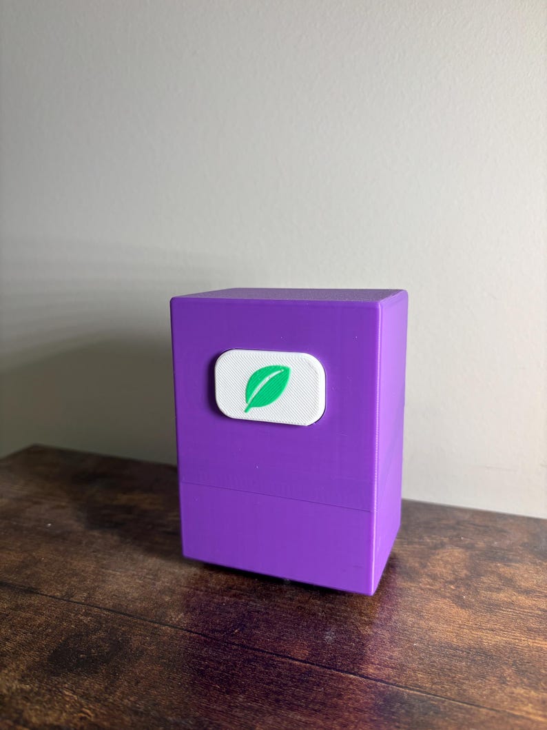 May include: A rectangular purple storage box with a smooth matte finish. Centered on the front face is a white, rounded rectangular emblem featuring a green leaf icon. The box has a horizontal seam line separating the lid from the base section.
