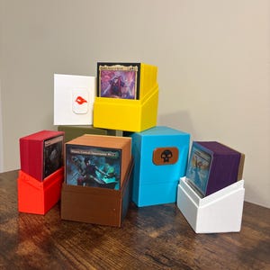 May include: A collection of six plastic trading card deck boxes in yellow, red, brown, blue, white, and olive green. Each box holds a stack of cards, with the front card visible. The blue box features a brown skull emblem, and the white box has a red icon.