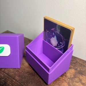 May include: A purple plastic deck box for trading cards with an open top and a detached lid. The lid features a white rectangular emblem with a green leaf icon. A stack of cards with a dark purple and blue circular graphic is inside the box.