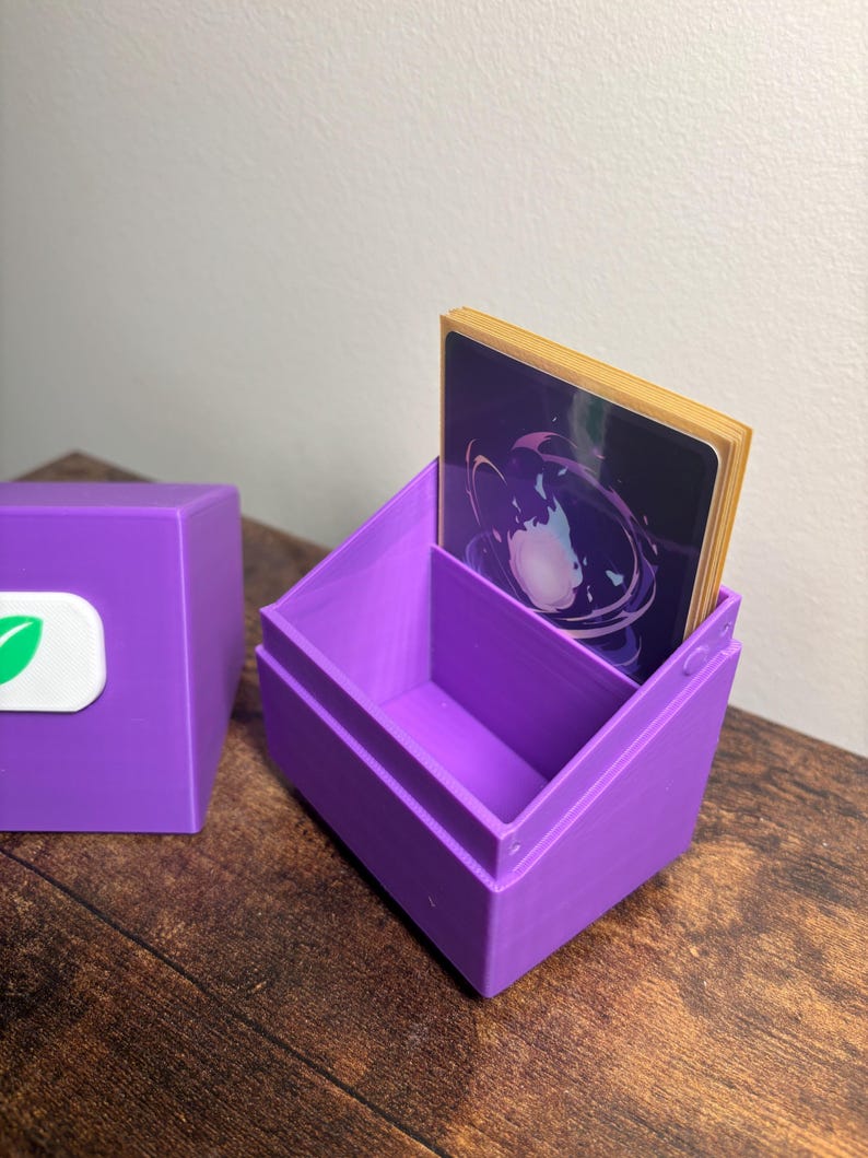 May include: A purple plastic deck box for trading cards with an open top and a detached lid. The lid features a white rectangular emblem with a green leaf icon. A stack of cards with a dark purple and blue circular graphic is inside the box.