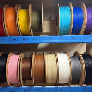 May include: A collection of 3D printer filament spools arranged on two wooden shelves. The spools feature a variety of colors including red, orange, yellow, green, lime, light blue, blue, purple, black, pink, bronze, brown, beige, white, and gray.