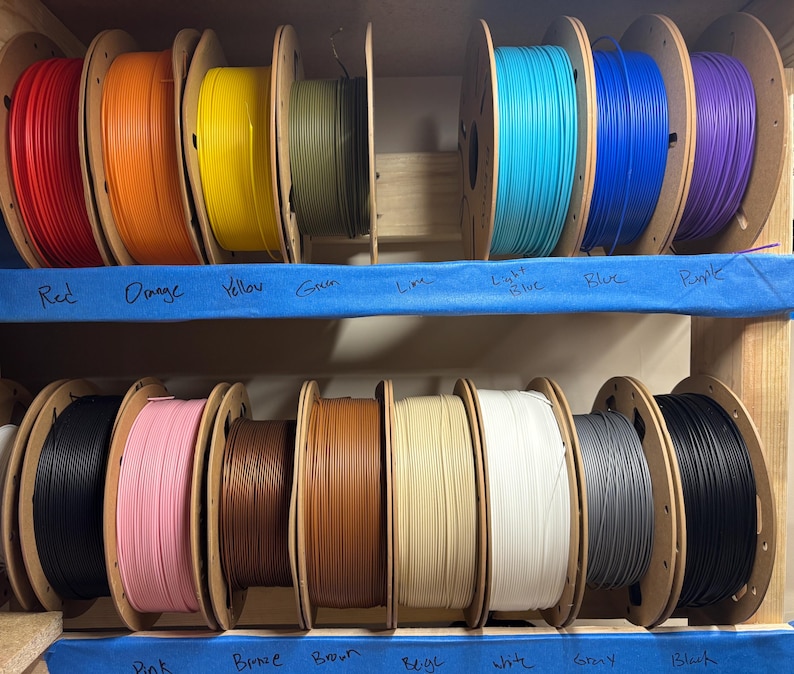 May include: A collection of 3D printer filament spools arranged on two wooden shelves. The spools feature a variety of colors including red, orange, yellow, green, lime, light blue, blue, purple, black, pink, bronze, brown, beige, white, and gray.
