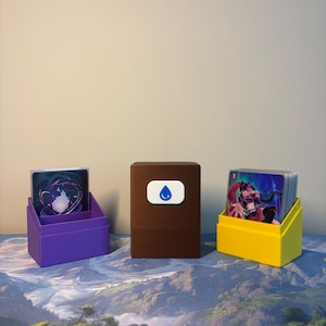May include: A brown rectangular deck box with a white emblem featuring a blue water drop icon, flanked by a purple card holder on the left and a yellow card holder on the right, each containing a stack of trading cards with colorful illustrations.