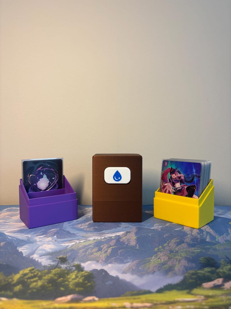 May include: A brown rectangular deck box with a white emblem featuring a blue water drop icon, flanked by a purple card holder on the left and a yellow card holder on the right, each containing a stack of trading cards with colorful illustrations.