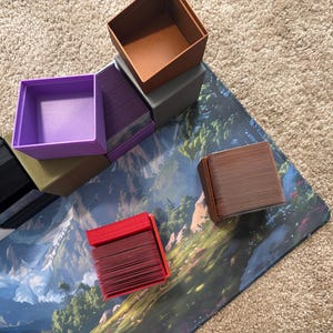 May include: A collection of square plastic card storage boxes in various solid colors including red, purple, black, brown, and grey. Some boxes are open and empty, while others are filled with stacks of playing cards, arranged on a surface.