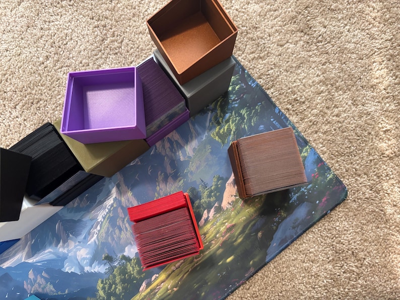 May include: A collection of square plastic card storage boxes in various solid colors including red, purple, black, brown, and grey. Some boxes are open and empty, while others are filled with stacks of playing cards, arranged on a surface.
