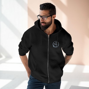 Puede incluir: Una sudadera negra con cremallera, capucha con cordones y cierre frontal plateado. En el pecho izquierdo, un logotipo circular muestra las letras WN rodeadas por un diseño de corona decorativa en tonos blancos y grises.