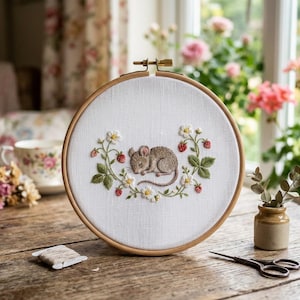 May include: A circular wooden embroidery hoop containing white fabric with a hand-stitched design of a small brown mouse sleeping in the center, surrounded by green leaves, white flowers, and small red strawberries arranged in a curved pattern.